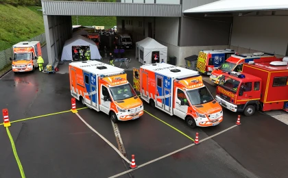 Mehrere Rettungsfahrzeuge, darunter Krankenwagen und Feuerwehrfahrzeuge, stehen auf einem asphaltierten Platz vor einer Überdachung. Im Hintergrund sind Zelte und Einsatzpersonal zu sehen.