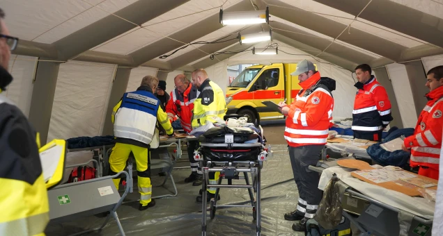 Ein Notfallzelt mit mehreren Personen in Einsatzkleidung, die sich um eine Trage und Tische versammelt haben. Im Hintergrund steht ein Rettungswagen.
