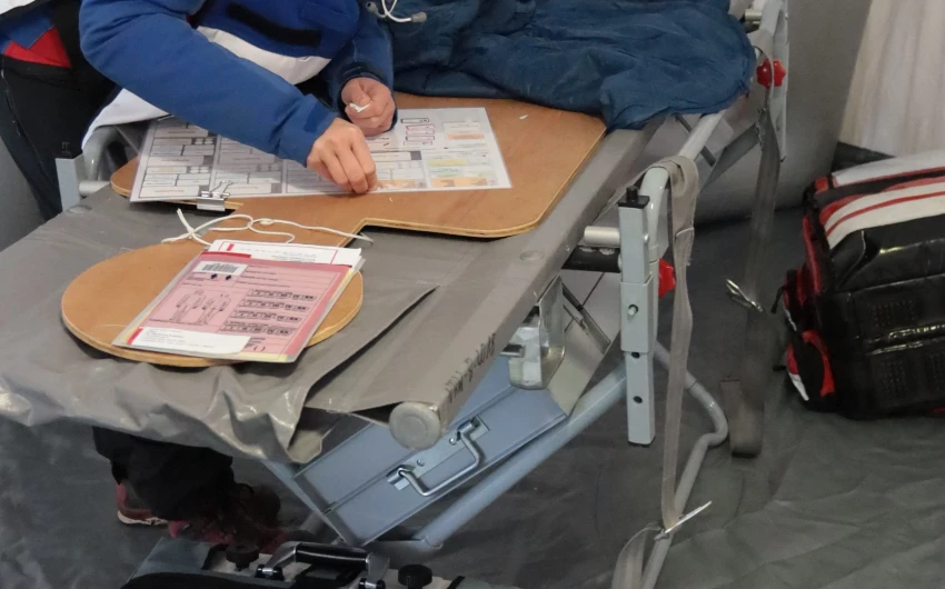 Eine Person in blauer Weste und rotem Pullover beugt sich über eine Trage mit einem Klemmbrett. Im Vordergrund eine Tasche mit der Aufschrift 'LIFE-BASE III'.