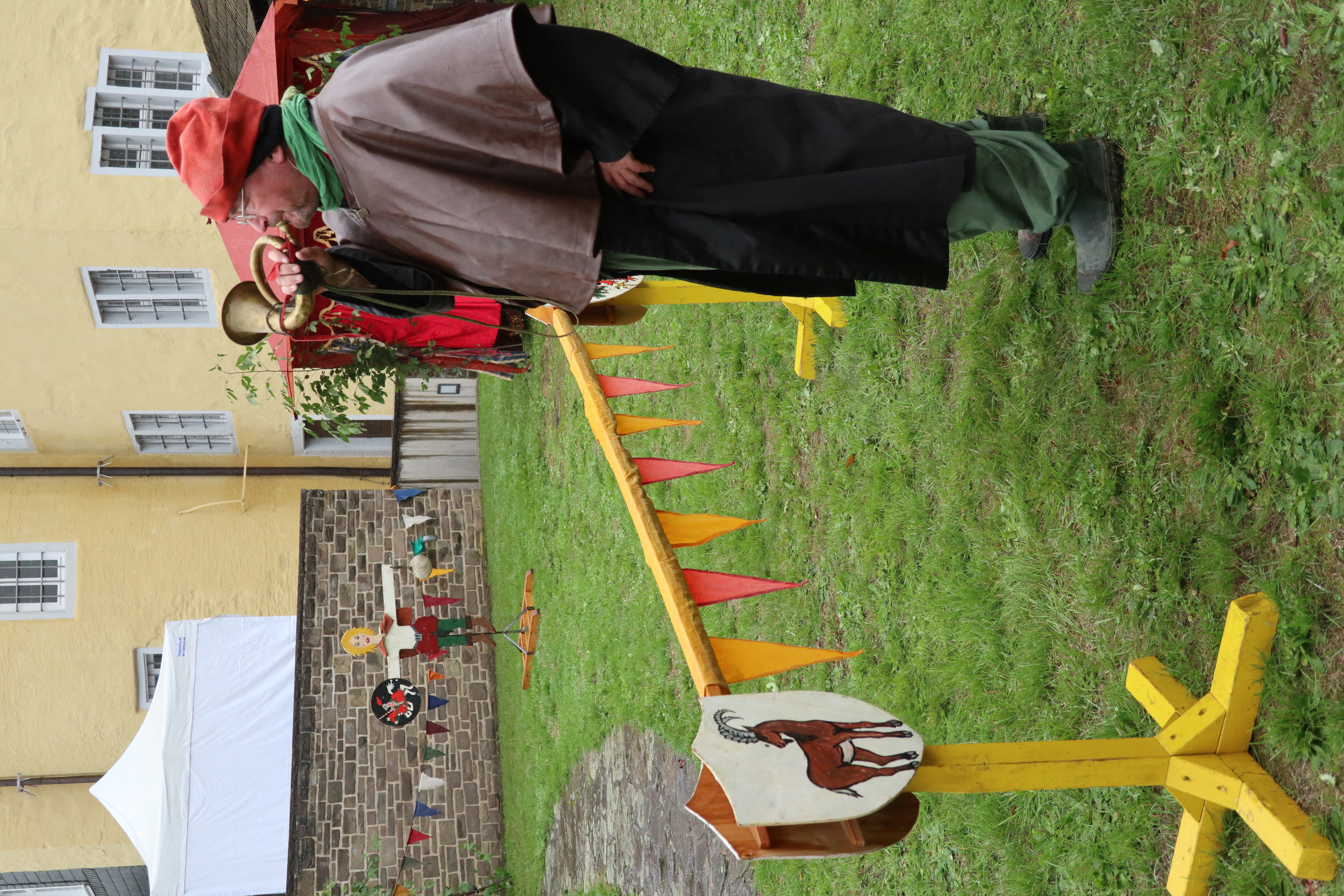 Ein Mann in mittelalterlicher Kleidung mit roter Mütze bläst ein Horn. Im Hintergrund stehen eine gelbe Hauswand, eine Zielscheibe und ein Pavillon.