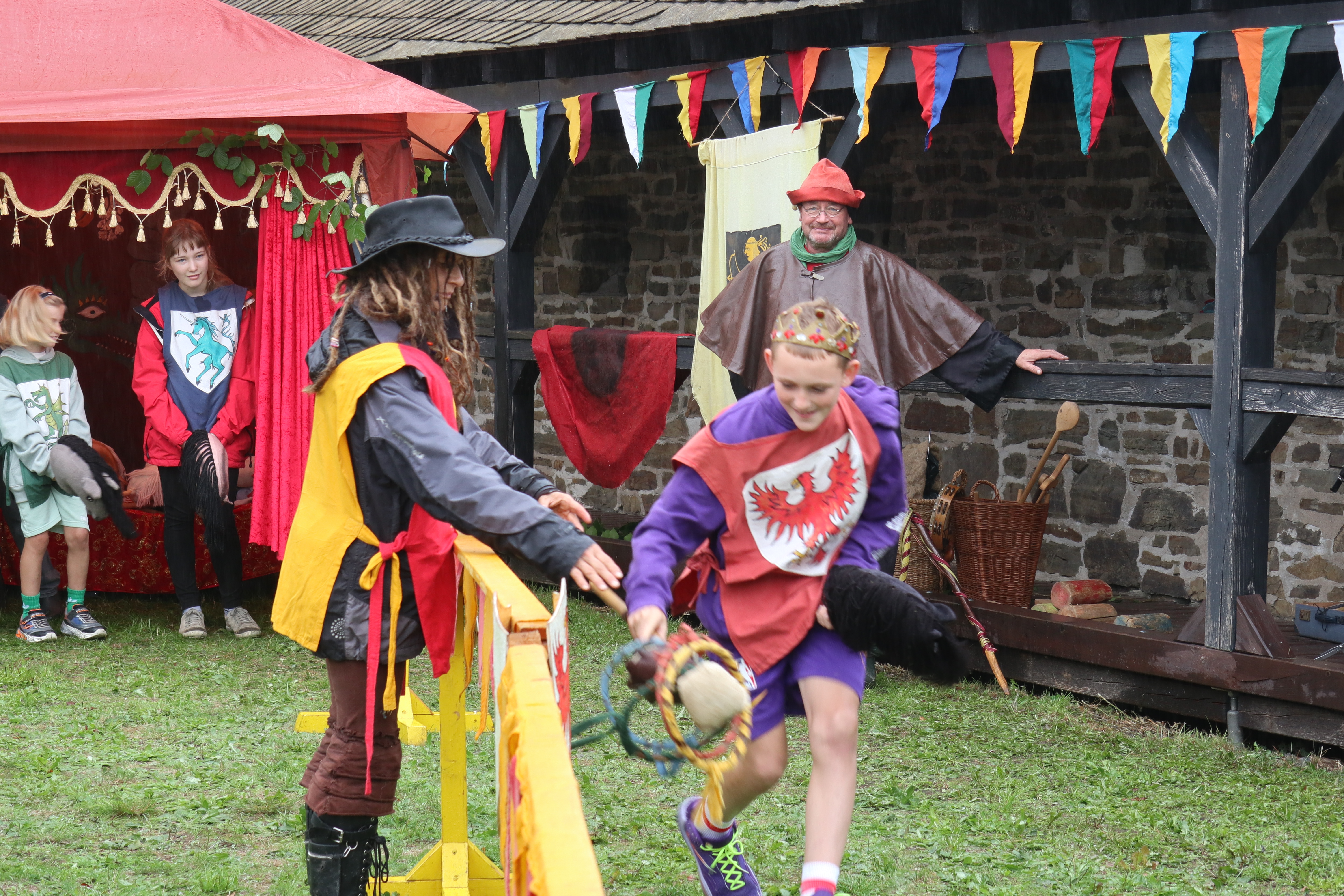 Kinder in mittelalterlichen Kostümen bei einem Festspiel. Ein Junge mit Krone und Wappen läuft, während andere Kinder und ein Erwachsener zusehen.