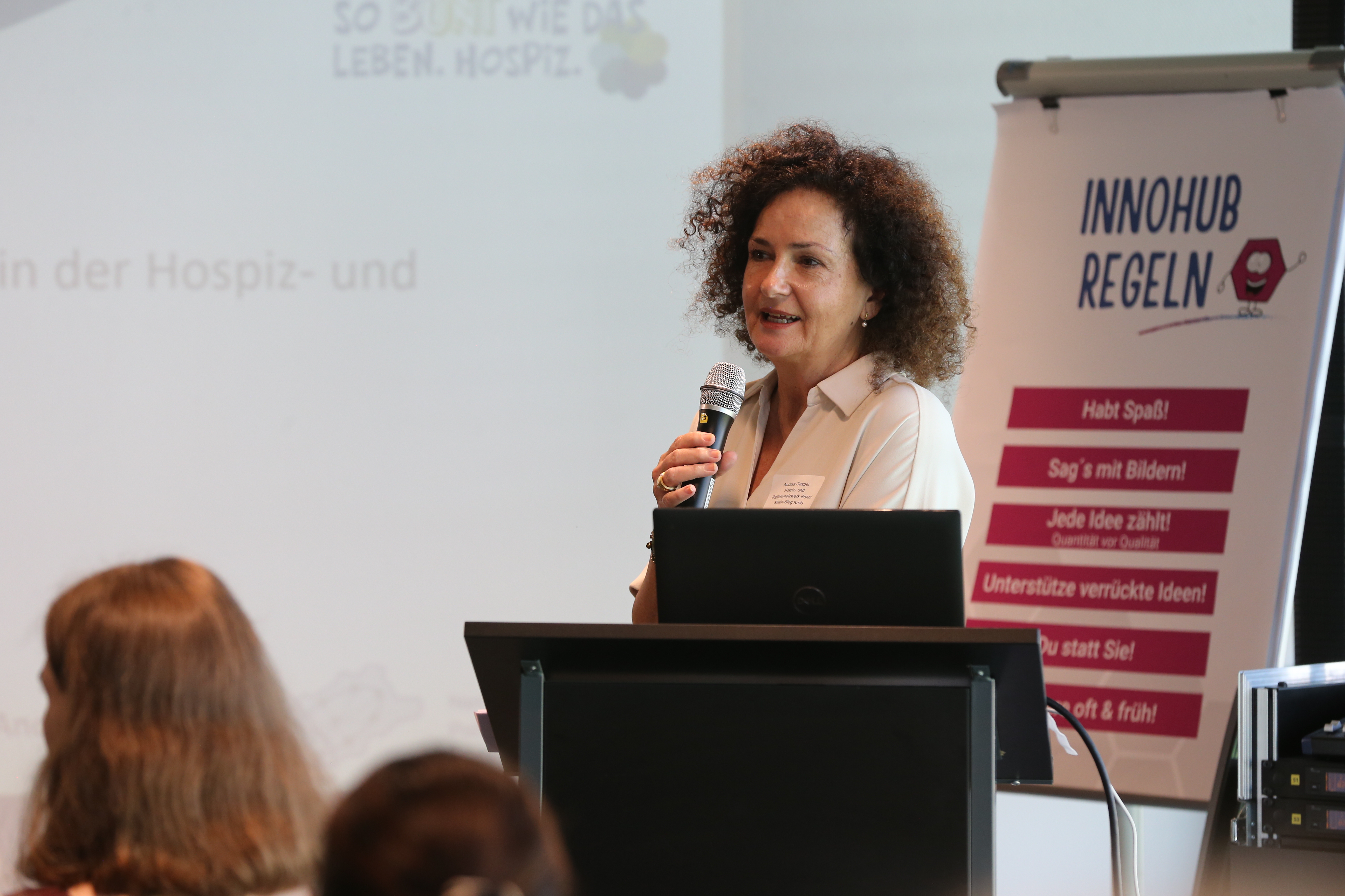 Eine Frau spricht mit einem Mikrofon vor einem Rednerpult. Im Hintergrund ist ein Plakat mit der Überschrift 'Innohub Regeln' und weiteren Texten sichtbar.